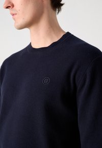 Navy blue knit sweater featuring a round neckline and a small embroidered logo on the chest, showcasing a smooth texture and fitted design.