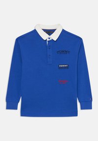 Polo bleu à manches longues avec col blanc, comportant des broderies "TOMMY HILFIGER" et des patchs de logo. Patte de boutonnage et détails au niveau des poignets.