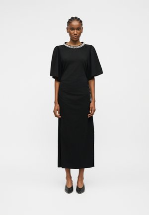 Mujer de pie vistiendo un vestido midi negro con mangas abullonadas y un escote adornado con joyas, combinado con zapatos negros de punta puntiaguda.