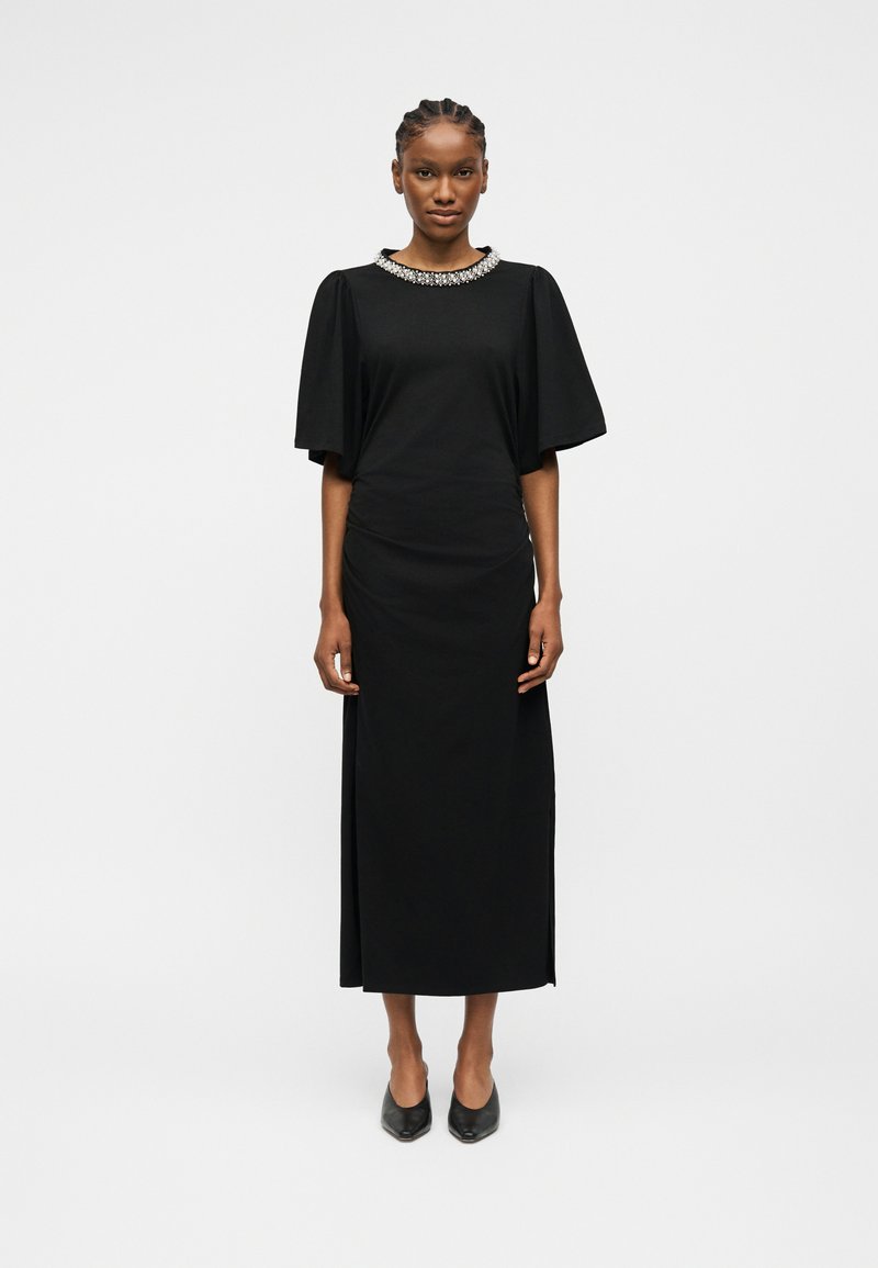 Γυναίκα που στέκεται φορώντας ένα μαύρο μίντι φόρεμα με φουσκωτά μανίκια και λαιμόκοψη διακοσμημένη με κοσμήματα, συνδυασμένο με μαύρες παπούτσια με μυτερή μύτη.