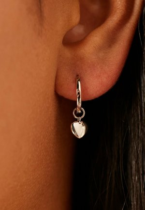 Close-up van een oor met een kleine zilveren creoolring met een hanger in de vorm van een hartje.