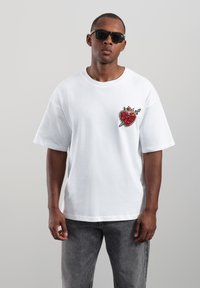 Weißes oversize T-Shirt mit einem strukturierten roten Herz- und Pfeil-Aufnäher auf der Brust. Weicher Baumwollstoff, Rundhalsausschnitt, getragen mit grauer Jeans.