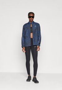 Blauwe, patroon sportjack met een hoge kraag, gecombineerd met een zwart crop-top en zwarte leggings. Afgewerkt met zwarte sportschoenen.