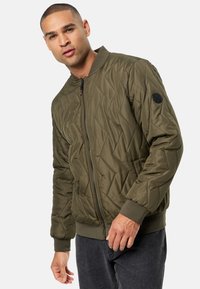 Chaqueta bomber acolchada en verde oliva con cierre de cremallera, puños acanalados y un parche en la manga izquierda. Diseñada con un patrón texturizado.