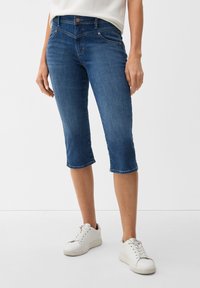 Dunkelblaue Denim Capri-Hosen mit mittelhohem Bund, Fünf-Taschen-Design und rohem Saum. Kombiniert mit weißen Sneakers.