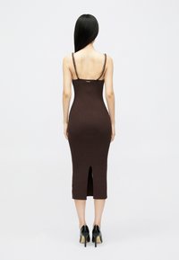 Καφέ ριμπ μίντι φόρεμα με σχισμή στην πλάτη, λεπτές τιράντες διακοσμημένες με αλυσίδες και κομψή γραμμή. Ορατές ψηλές γόβες από κάτω.