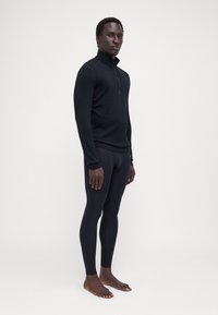 Schwarzes Langarmoberteil mit Reißverschlusskragen und enganliegenden schwarzen Leggings, beide aus glattem, elastischem Material. Barfußhaltung vor einem hellen Hintergrund.