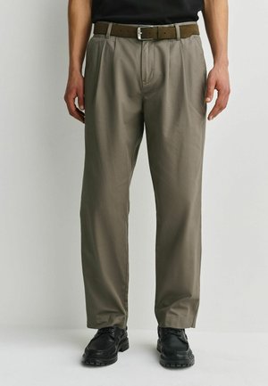 Mann trägt khakifarbene, plissierte Hose mit braunem Gürtel und schwarzen Schnürschuhen, steht vor einem schlichten hellen Hintergrund.