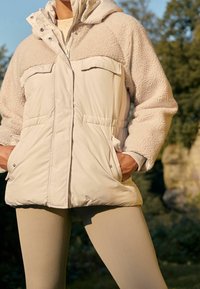 Beige pufferjack met een fuzzy, lichtbruin bovenstuk en twee voortassen. Gecombineerd met lichte geribbelde leggings.