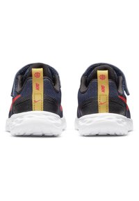 Nike sportbačiai iš tamsiai mėlyno tinklinio audinio su juodais akcentais, raudona swoosh logotipu ir geltonu atramos kilpelė. Balta tekstūruota padas. Uždara nugarinė dalis, sportiškas dizainas.