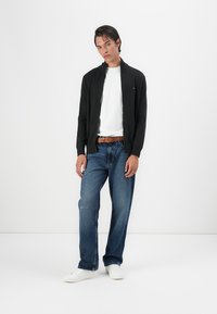 Crna zip-up cardigan preko bijele majice, uparen s svijetloplavim trapericama i bijelim tenisicama. Vidljiv smeđi pojas; opušteni kroj; minimalistički dizajn.
