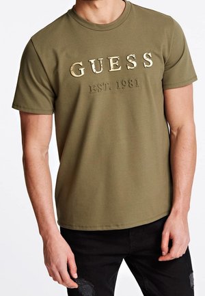 Mężczyzna w oliwkowozielonym T-shircie z krótkim rękawem z wytłoczonym napisem „GUESS EST. 1981” z przodu, w połączeniu z czarnymi, podartymi jeansami.