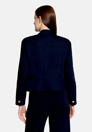 Vrouw met lang bruin haar draagt een getailleerd donkerblauw jasje met knoopmanchetten, met haar rug naar de kijker tegen een effen lichte achtergrond.