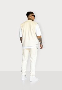 Cremefarbener ärmelloser Top über einem weißen T-Shirt, kombiniert mit weißen Jogginghosen und Turnschuhen. Hat eine glatte Textur, ein lässiges Design und minimale Akzente.