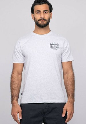 Homme aux cheveux foncés et à la barbe portant un t-shirt gris clair avec un motif de voiture d'Indianapolis et un pantalon noir, debout devant un fond uni.