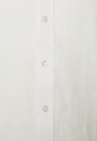 Camisa de lino blanca con un tapón de botones que presenta botones de plástico transparente. Textura suave y patrón de tejido sutil visible en la tela.