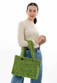 Grüne, strukturierte Tragetasche mit zwei stabilen Griffen, rechteckiger Form und einem runden Hardware-Akzent. Getragen mit einem hellbeigen Oberteil und blauen Jeans.