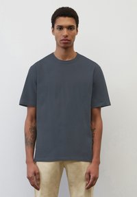 Czarnoszary krótki rękaw T-shirt z okrągłym dekoltem, wykonany z gładkiego materiału, noszony z jasnymi spodniami, z widocznymi tatuażami na ramionach.