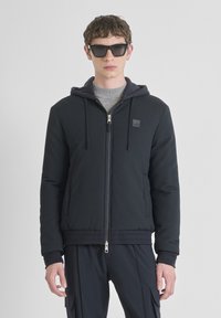 Een jonge man draagt een donkergequilted ritshoodie met trekkoorden, een grijs shirt eronder, zwarte cargobroek en grote zwarte zonnebril, staand.