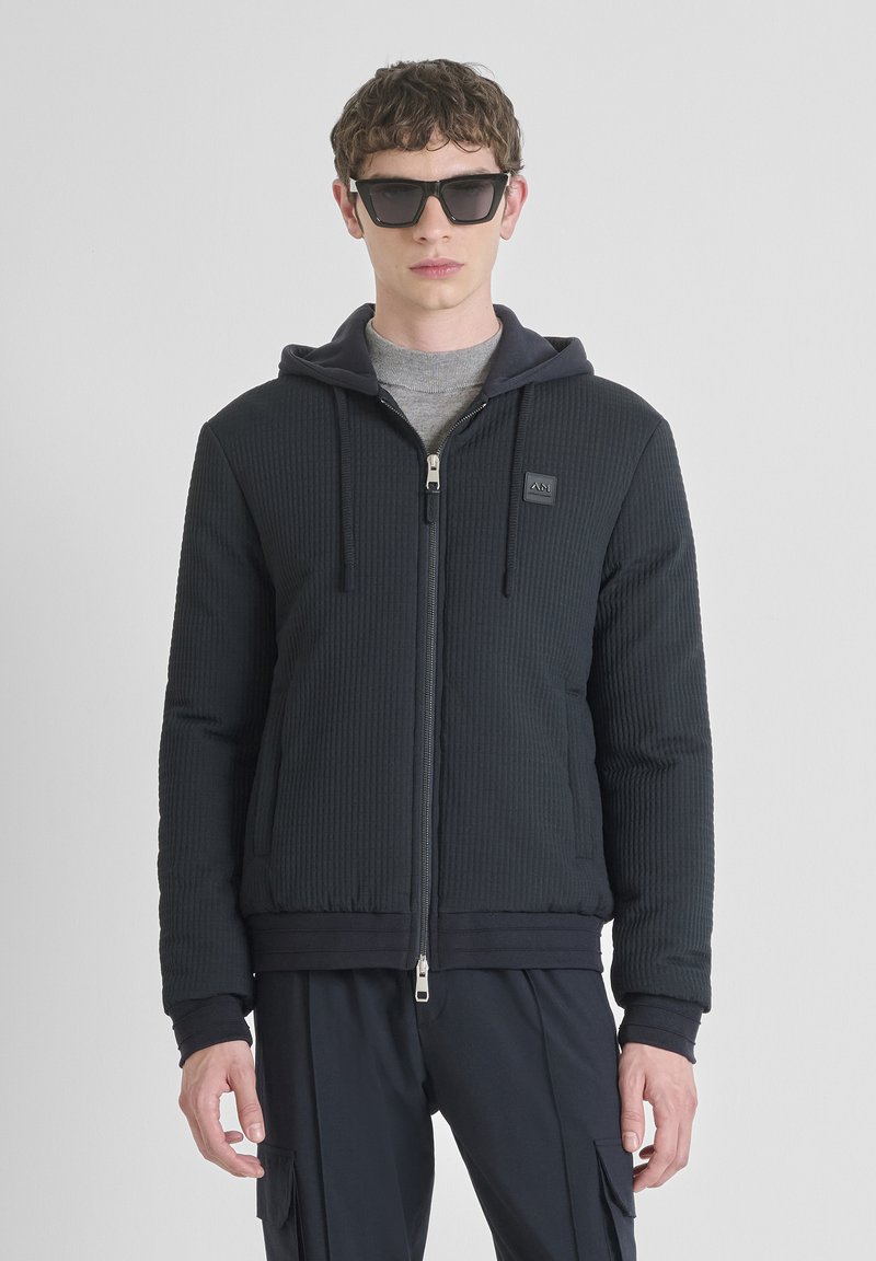 Een jonge man draagt een donkergequilted ritshoodie met trekkoorden, een grijs shirt eronder, zwarte cargobroek en grote zwarte zonnebril, staand.