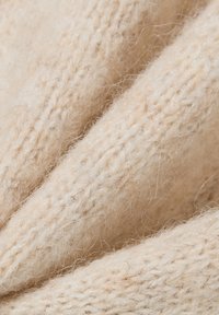 Tessuto lavorato a maglia beige con una consistenza morbida e pelosa. Presenta cuciture visibili e pieghe delicate, creando un aspetto caldo e accogliente.