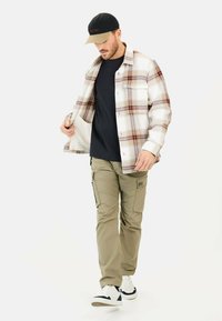 Geruite flanellen overhemd met rode en bruine accenten, zwart T-shirt, kaki cargo pants, witte sneakers en een zwarte pet met lichte rand.