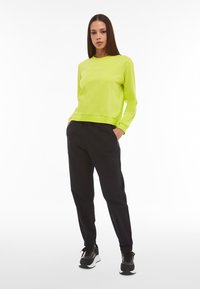 Neon-grüner Pullover mit Rundhalsausschnitt und gerippten Bündchen, kombiniert mit schwarzen, schmal zulaufenden Hosen und schwarzen Sneakers mit weißen Sohlen.