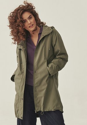 Kobieta o kręconych włosach w zielonej parce z kapturem, nałożonej na fioletowy sweter i ciemne spodnie, pozująca z rękami w kieszeniach.