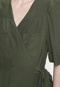 Olive green wrap top made of textured fabric, featuring short sleeves and two front pockets. Includes a tie at the waist for closure.