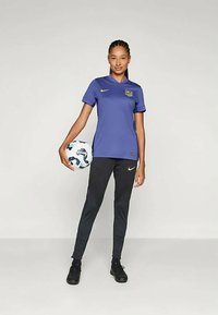 Lilla fotballsdrakt med en gul logo, kombinert med svarte bukser med et strukturert design. Modellen holder en mønstrete fotball.