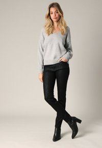 Pull gris à col en V avec des poignets côtelés, associé à un pantalon noir texturé présentant un motif subtil. Des bottines noires à chevilles complètent la tenue.