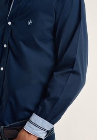 Camisa de botones azul marino con una textura suave, botones blancos y un puño azul claro con rayas marinas. Presenta un logo bordado en el pecho.