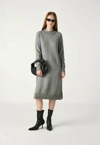 Kvinne iført en grå langt-sleeve strikkekjole, svarte hælhøye støvler, solbriller, og holder en svart håndveske mot en enkel lys bakgrunn.