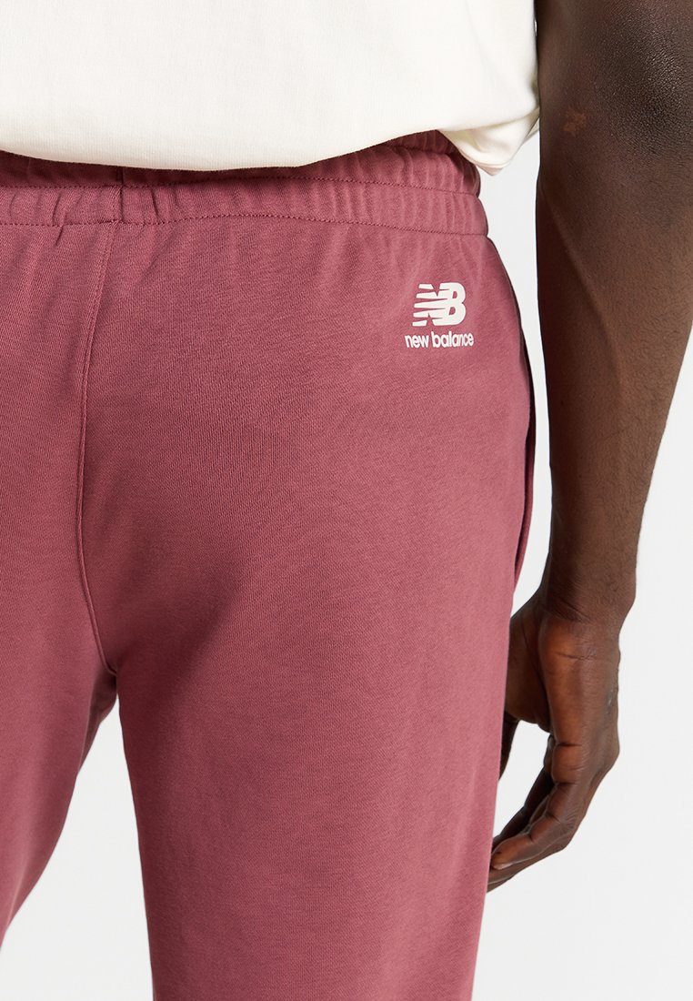 Person, der maroonfarbene New Balance Jogginghosen mit elastischem Bund und weißem Logo am Oberschenkel trägt, steht vor einem schlichten Hintergrund.