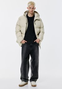 Helle beige Daunenjacke mit Kapuze, schwarzer Kabelstrickpullover, übergroße dunkelblaue Jeans und weiße Sneakers mit beigen Sohlen.