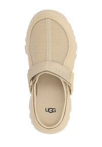 Zapato slip-on de red en color beige con suela de goma, diseño estructurado y correa ajustable. Cuenta con material transpirable y la marca UGG en la plantilla.