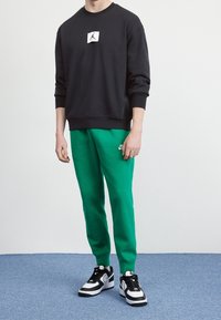 Sudadera negra con parche del logo de Jordan, combinada con pantalones de chándal verde vibrante que presentan un logo de Nike, y zapatillas negras y blancas.
