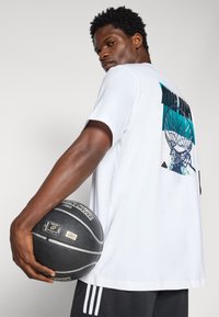 Bílý tričko s krátkým rukávem s barevným grafickým designem basketbalové sítě na zádech. Model drží černý basketbal.