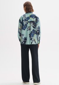 Camicetta floreale azzurro chiaro con motivi blu scuro e bianchi, vestibilità ampia, dettagli con bottoni ai polsini, abbinata a pantaloni larghi neri.