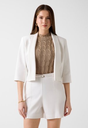 Femme portant un blazer blanc sur mesure avec manches aux coudes, haut beige en maille torsadée, shorts blancs ceinturés, boucles d'oreilles créoles dorées et bracelet.