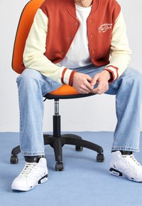 Röd och krämfärgad varsityjacka med tryckknappar, vit t-shirt, ljusblå jeans och vita sneakers. Sitter på en orange kontorsstol.