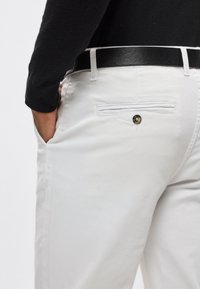 Białe bawełniane spodnie z tylną kieszenią zapinaną na guzik oraz czarnym skórzanym paskiem. Krój cechuje się smukłą, dopasowaną sylwetką.