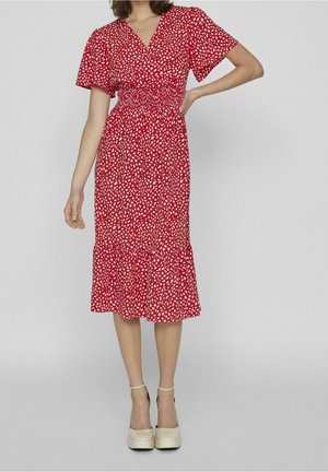 Vrouw draagt een rode midi-jurk met witte onregelmatige stippen, korte mouwen, een taille met smockwerk en een versierde zoom, gecombineerd met crèmekleurige plateauhakken.