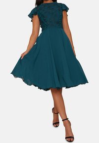 Turkos A-linje klänning med korta volangärmar och spetstopp. Flödande tyg med en fåll som faller under knäna. Svarta sandaler med remmar.