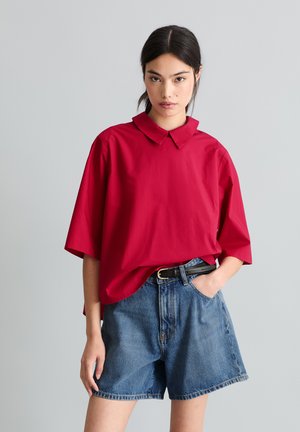Junge Frau trägt ein locker sitzendes rotes Hemd mit Kragen und hoch taillierte Jeansshorts mit schwarzem Gürtel, steht vor einem schlichten grauen Hintergrund.