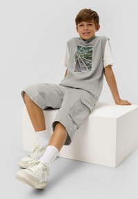 Szara koszulka bez rękawów z graficznym nadrukiem, zestawiona z jasnoszarymi szortami cargo. Białe sneakersy i skarpetki dopełniają strój.
