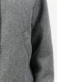 Giacca grigia in feltro caratterizzata da una texture morbida, una vestibilità rilassata e un design minimalista. Include una tasca laterale e cuciture per la struttura.