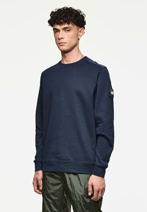 Hombre con cabello rizado que lleva una sudadera de manga larga azul marino y pantalones brillantes verde oliva, de pie contra un fondo blanco liso.