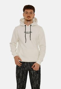 Light cream hoodie with black drawstrings and front pocket, featuring "Picaldi Jeans Berlin" text in black. Paired with patterned dark pants.