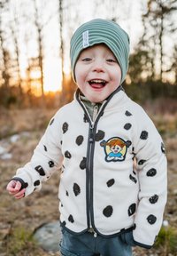 Fleece-takki, jossa on mustat pilkut valkoisella, vetoketjulla edessä ja harmaalla reunuksella. Sisältää kirjotun patchin hahmosta. Vihreä raidallinen pipot.